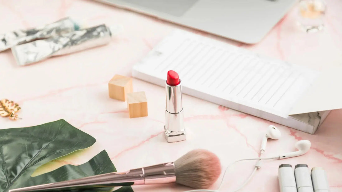 A silver lipstick with a bright red bullet stands on a pink marble surface, surrounded by beauty and lifestyle props including a makeup brush, white earbuds, a notebook, and small wooden blocks.