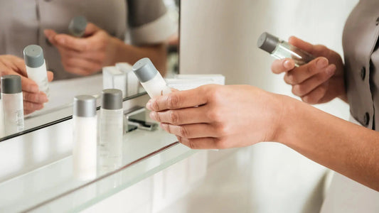 A person holding a small cosmetic bottle in front of a mirror, with several similar bottles arranged on a glass shelf in a bright bathroom or vanity setting.